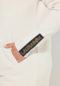 Lichtcrème kleurig sweatshirt met ribgebreide manchetten. De linkermanchet heeft een marineblauwe band met gele tekst "LAURASON". De voorkant heeft een zijzak.