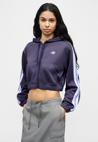 Νεαρή γυναίκα με μακριά σκούρα μαλλιά, φορώντας κοντό μπλε σκούρο φούτερ με λευκές ρίγες και γκρι φούστα, όρθια με τα χέρια στις τσέπες.