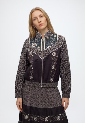 Femme aux cheveux blonds portant une veste noire zippée à motifs et une jupe fleurie assortie, debout devant un fond blanc uni.