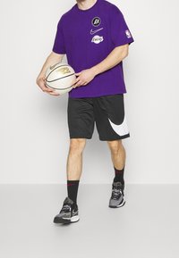 Camiseta morada de Nike con logos, pantalones cortos negros con el Swoosh blanco y zapatos negros con detalles en gris; sosteniendo un balón de baloncesto.