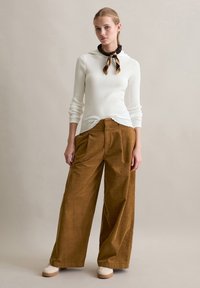 Witte geribde longsleeve top met een capuchon, gecombineerd met brede bruine corduroy broek. Model draagt een gestructureerde sjaal en lichte schoenen.