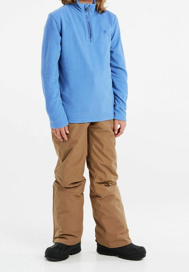 Jasnoniebieski polarowy sweter z ćwierć zamkiem i logo, w połączeniu z beżowymi spodniami narciarskimi i czarnymi butami. Fleece o fakturze, luźny krój i wzmocnione szwy.