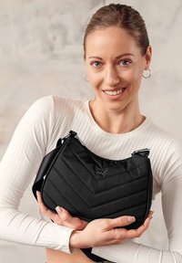 Černá prošívaná kabelka přes rameno s žakárovým vzorem, má zakřivený vršek a nastavitelný popruh, kterou drží osoba v světlejším topu.