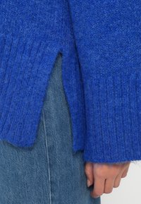 Μπλε πλεκτή μπλούζα με ριγέ υφή και πλάγιο άνοιγμα. Μαλακό υλικό, ελαφρώς φαρδιά εφαρμογή και μακριά μανίκια. Συνδυασμένη με τζιν.