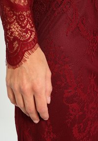 Burgundy lace dress with textured floral patterns, featuring intricate lace sleeves and fringing detail at the cuff. Smooth underlying fabric.