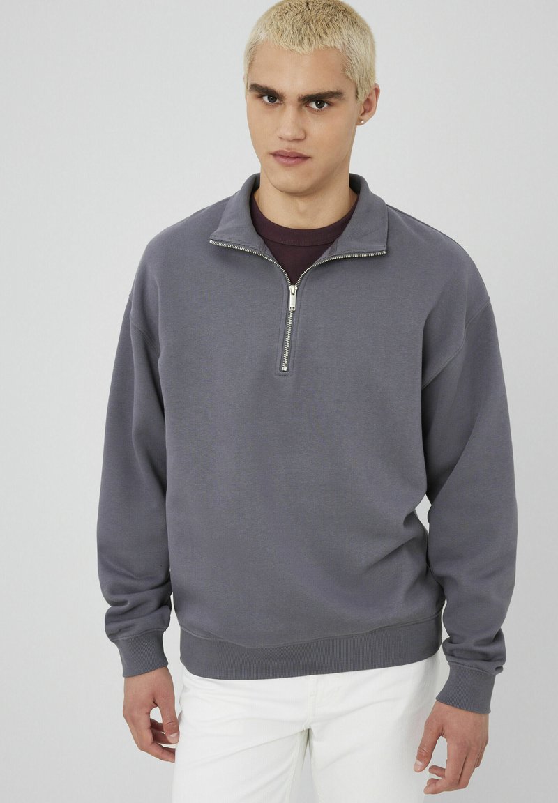 Sweatshirt zippé gris avec un col haut, coupe décontractée, poignets et ourlet côtelés. Porté par-dessus une couche violet foncé, associé à un pantalon blanc.