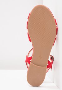 Punainen mokkabootsikorko, jossa on litteä, tekstuurinen beige pohja ja säädettävä hihna. Pohjassa on ilmoitettu koko 37. Takana suljettu ja sivu avonainen muotoilu.