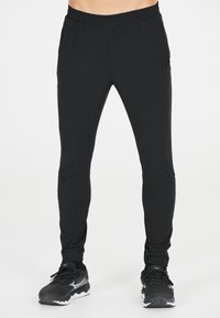 Zwarte sportbroek met een elastische tailleband, taps toelopende broekspijpen, twee zijzakken en een gladde, lichte stof. Gecombineerd met zwarte hardloopschoenen.