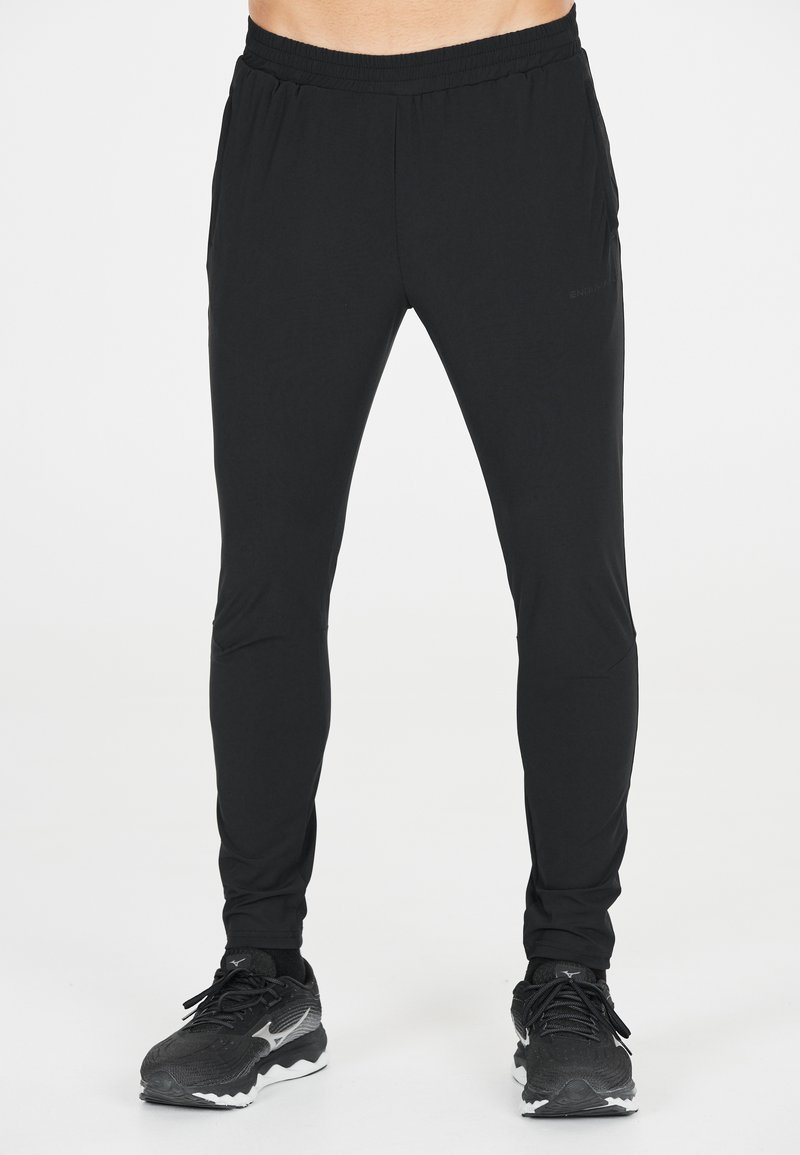 Zwarte sportbroek met een elastische tailleband, taps toelopende broekspijpen, twee zijzakken en een gladde, lichte stof. Gecombineerd met zwarte hardloopschoenen.