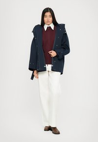 Navy waterproof jacket with a hood, fastened by snaps, worn over a burgundy knit sweater and white wide-leg pants, paired with brown boots.