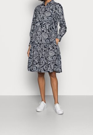 Woman wearing a black and white paisley patterned long-sleeve dress with collar, paired with white sneakers, standing against a plain background.