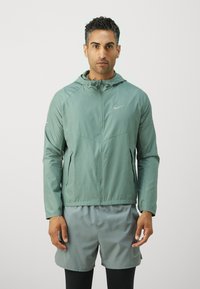 Veste zippée vert clair avec capuche, arborant un logo Nike, associée à un short gris. Matériau lisse et léger, design sportif.