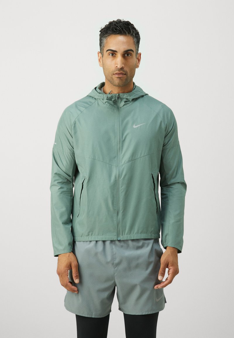 Světle zelená zipová bunda s kapucí, se znakem Nike, v kombinaci s šedými šortkami. Hladký, lehký materiál, sportovní design.
