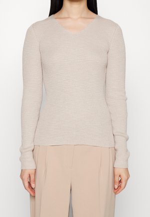 Kobieta ubrana w beżowy prążkowany sweter z długim rękawem i dekoltem w serek, wsunięty w jasnobeżowe, wysokowalne spodnie z plisami.