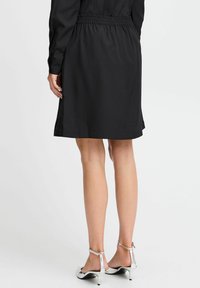 Schwarzes knielanges Kleid mit elastischem Bund, glatter Textur und kurzen Ärmeln. Paar silberne Absatzsandalen sind an den Füßen sichtbar.