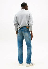 Felpa grigia, jeans blu con texture sbiadita e sneakers bianche. Dettagli di cucitura visibili e patch di pelle rettangolare sui jeans.