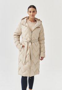 Beige quilted puffer coat with a hood, tie belt, and chevron pattern. Features a front zipper and side pockets, designed for warmth.