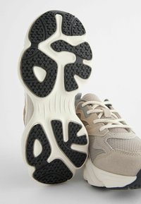 Sko med en beige mesh-overdel, detaljeret med ruskindselementer, og en hvid gummisål med et mønstret sort tread for godt greb.