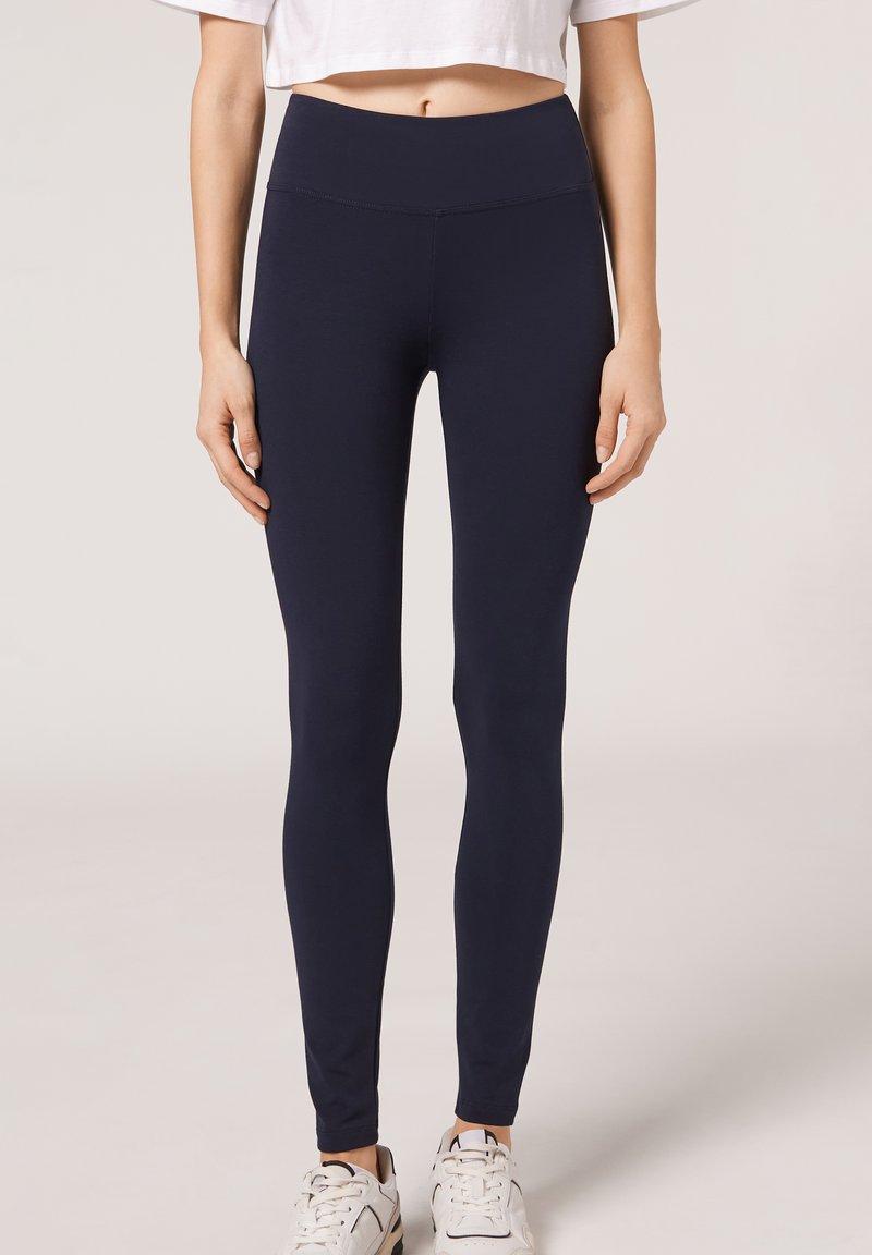 Leggings bleu marine en tissu extensible, taille haute, texture lisse, silhouette ajustée, associés à des baskets blanches.