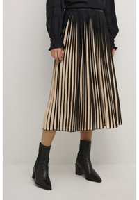 Woman wearing a black long-sleeve top, beige and black vertical striped pleated midi skirt, and black ankle boots with block heels.