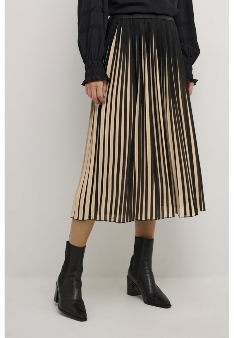 Woman wearing a black long-sleeve top, beige and black vertical striped pleated midi skirt, and black ankle boots with block heels.