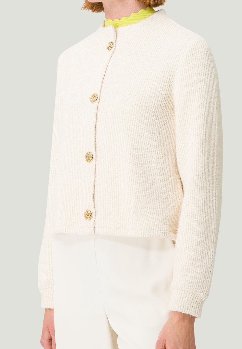 Cardigan crema con texture e design corto, caratterizzato da bottoni decorativi dorati e scollo tondo. Indossato sopra una maglietta verde brillante.