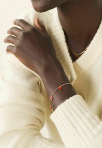 Gold bracelet with red heart charms, worn on a dark arm. The wrist is adorned with a cream knitted sweater, featuring ribbed texture.