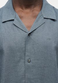 Άνδρας που φοράει ανοιχτό μπλε πουκάμισο με κουμπιά και διακριτικό λογότυπο "cK" στο στήθος.