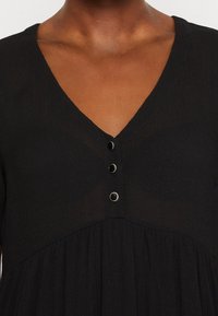 Vestido negro con escote en V, que presenta una tapeta con botones y una falda fruncida. La tela tiene una apariencia texturizada y ligeramente transparente.