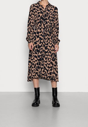 Kvinde iført en langærmet, læg-lang kjole med leopardmønster og taljebånd, kombineret med sorte ankelstøvler, der står på en grå gulv.
