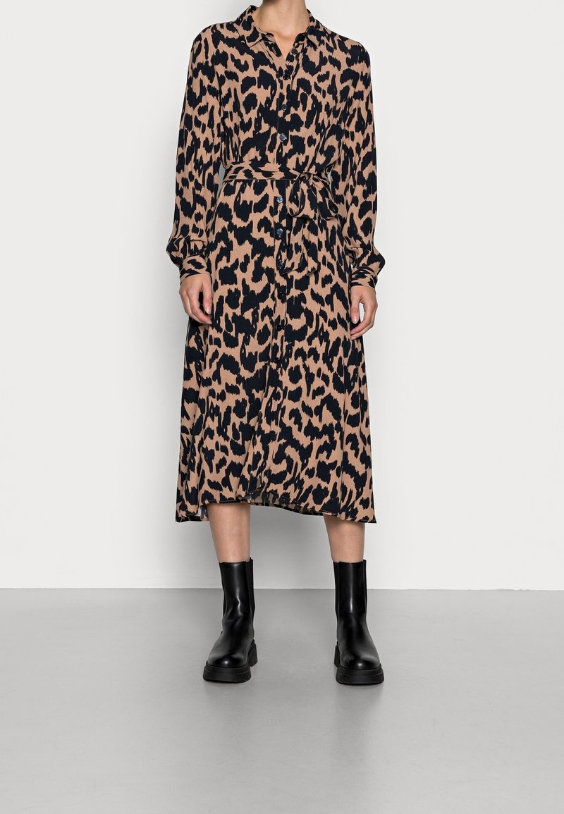 Femme portant une robe à manches longues, longueur mollet, imprimé léopard avec une ceinture à la taille, associée à des bottines noires, debout sur un sol gris.
