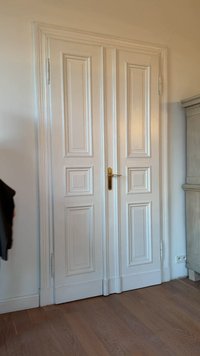 Fehér duplaajtók függőleges panelekkel, arany színű fogantyúkkal és sima felülettel, világos belső fal és fa padló előtt.