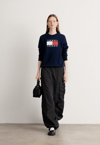 Felpa girocollo navy con logo "TOMMY JEANS" in lettere rosse e bianche. Abbinata a pantaloni cargo neri larghi e accessori neri.