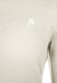 Ljusgrå stickad tröja med rund halsringning och en liten, diskret logga broderad på vänstra bröstområdet.