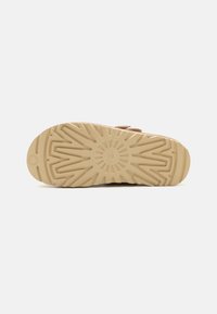 Tan platform shoe with a textured rubber outsole featuring a sunburst pattern and curved grooves for traction. Adjustable strap detail present.