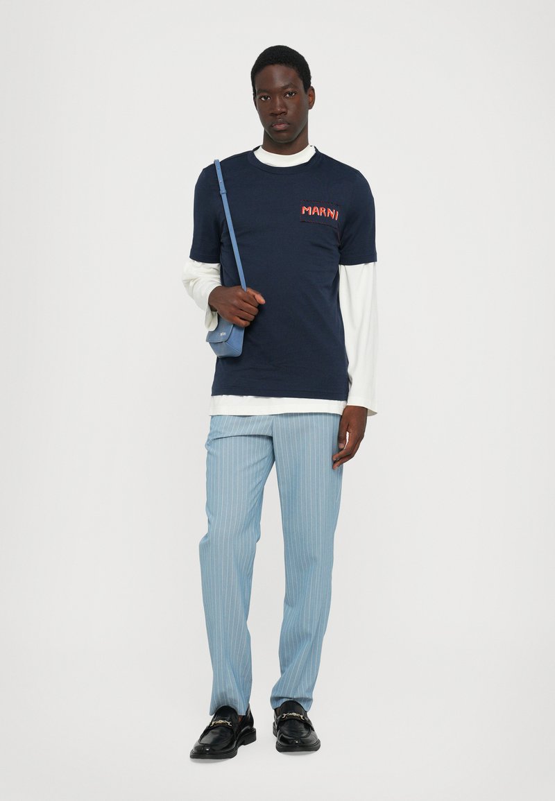 Camiseta de algodón azul marino con el logo rojo "MARNI", superpuesta sobre una camisa de manga larga blanca. Pantalones de rayas azul claro y mocasines negros, con un bolso de hombro azul.