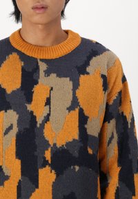 Strikkegenser med en rund oransje hals, med et fargeblokkert design i marineblått, oransje og beige med varierte teksturer og mønstre.