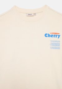 Krém színű, organikus pamut pulóver, kék grafikával, amelyen a "Cherry" szó szerepel. Kerek nyakú és texturált anyagú.