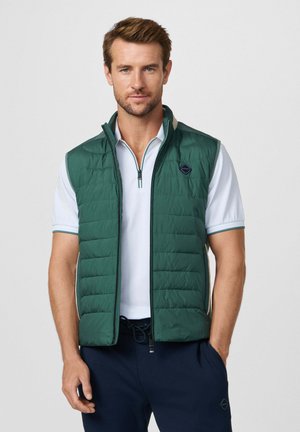 Homme portant un gilet matelassé vert sur un polo blanc à manches courtes avec fermeture éclair, et un pantalon bleu marine, debout avec une main dans la poche.