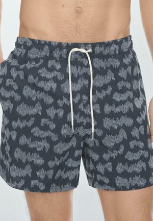 Torso masculin et parties supérieures des jambes portant un short de bain gris foncé avec des rayures blanches irrégulières et abstraites, et un cordon de serrage blanc noué à la taille.