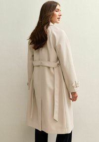 Trench beige con cintura de cinturón, hombreras y detalles de puño abotonado. Textura suave y diseño hasta la rodilla.