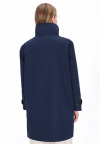 Mørkeblå frakke med høj krave, knappede manchetter og lige snit. Glat tekstur med minimal detaljering, designet til en skræddersyet pasform.