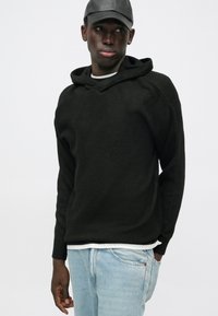 Black textured hoodie with a V-neck design, featuring a soft knit fabric and a relaxed fit. Worn over a white layered shirt and blue jeans.