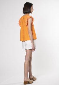 Top in lino arancione con dettagli in pizzo arricciato, abbinato a pantaloni corti in lino bianchi. Il modello indossa zoccoli beige con tacchi in legno.