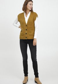 Senfgelbes Strickweste mit V-Ausschnitt, vier schwarzen Knöpfen und zwei Vorderknienten, kombiniert mit schwarzen schmalen Jeans und beigefarbenen Ankle Boots.