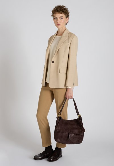 Beżowa marynarka o dopasowanym kroju z jednym guzikiem i kieszeniami z przodu, zestawiona z dopasowanymi, wąskimi spodniami. Brązowa pleciona torebka z złotym akcentem.