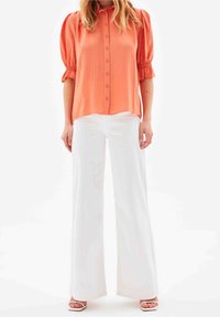 Blouse corail à boutons avec manches courtes bouffantes, présentant des rayures verticales, associée à un pantalon blanc à jambes larges et des sandales blanches à talons.