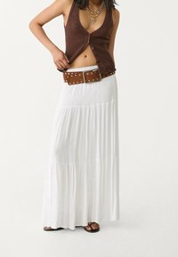 Brązowa dzianinowa kamizelka z dekoltem w serek, zestawiona z białą, warstwową maxi spódnicą. Dodatki to brązowy pasek z srebrnymi ćwiekami oraz płaskie sandały.