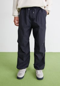 Pantalons noirs en nylon léger avec une taille élastique, poches latérales et une coupe décontractée, associés à des baskets grises sur un sol vert.