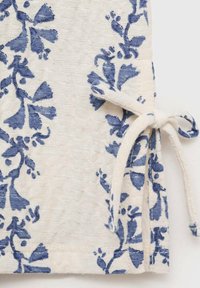 Tissu blanc avec un motif floral bleu, surface texturée, bords finis et détail noué. Présente de grands motifs de fleurs et de feuilles stylisés.